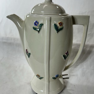 May include: A cream-colored ceramic electric coffee percolator featuring a ribbed lid, a side handle, and a spout. The body is decorated with small, raised, hand-painted floral motifs in blue, yellow, and purple with green leaves.