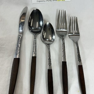 May include: A 5-piece Interpur stainless steel place setting from Japan featuring brown synthetic handles with a heart design. The set includes one knife, two spoons, and two forks. A label reads: Interpur Stainless Japan 5pc Place Setting Brown Synthetic Handle Heart Design 2 spoons, 2 forks, knife LOT 101.