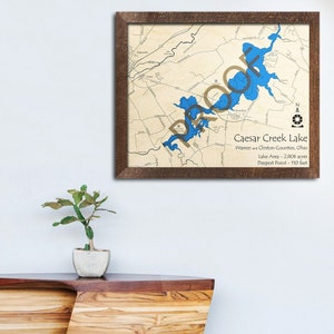 May include: A framed map of Caesar Creek Lake in Warren and Clinton Counties, Ohio. The map shows the lake's shape and surrounding area. The text below the map reads "Caesar Creek Lake, Warren and Clinton Counties, Ohio, Lake Area - 2,806 acres, Deepest Point - 110 feet."