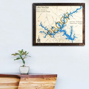May include: A framed map of Lake Sinclair in Georgia, with the lake's outline highlighted in blue. The map includes the lake's name, location, and size in acres. The word "PROUD" is written in gold letters over the lake.