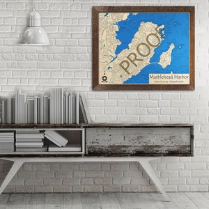 May include: A framed map of Marblehead Harbor, Essex County, Massachusetts. The map is printed on a cream-colored background with blue water features. The frame is brown wood.