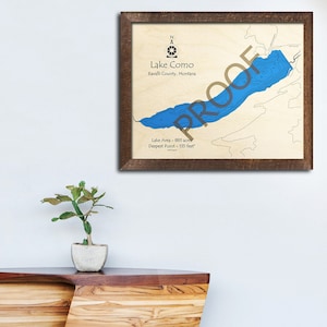 May include: A framed map of Lake Como in Ravalli County, Montana. The map shows the lake's shape and depth contours in blue. The text "Lake Como", "Ravalli County, Montana", "Lake Area - 883 acres", and "Deepest Point - 155 feet" is printed on the map.