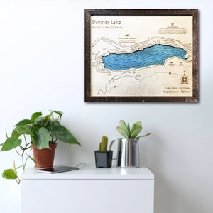 May include: A framed wooden map of Donner Lake in Nevada County, California. The map shows the lake's depth contours in blue, with surrounding land features in brown. The text "Donner Lake" and "Nevada County, California" is printed at the top of the map. The text "Lake Area - 840 acres" and "Deepest Point - 528 feet" is printed at the bottom of the map.