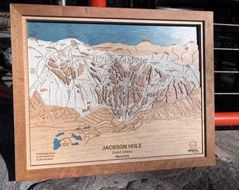 May include: A framed wooden map of Jackson Hole, Grand Tetons, Wyoming. The map shows the ski runs and terrain of the mountain. The map is surrounded by a light brown wooden frame.