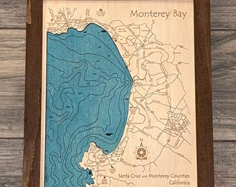 May include: A framed wooden map of Monterey Bay, California, with blue water and brown land features. The map includes the text "Monterey Bay" and "Santa Cruz and Monterey Counties, California".