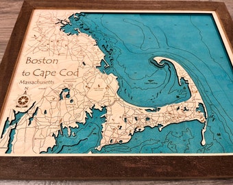 May include: A wooden map of Boston to Cape Cod, Massachusetts, with a blue water feature and brown wood for the land. The map is framed in a dark brown wood frame.