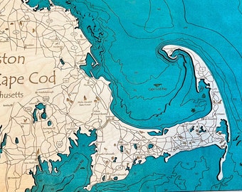 May include: A wooden map of Boston to Cape Cod, Massachusetts. The map is laser cut and features a blue ocean with depth lines and a beige landmass with roads and towns. The title "Boston to Cape Cod, Massachusetts" is written in brown text at the top of the map.