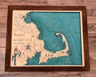 May include: A framed wooden map of the Boston to Cape Cod area in Massachusetts. The map is a 3D relief map with blue water and brown land. The map is framed in a dark brown wood frame.