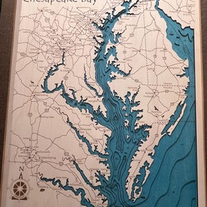 May include: A wooden map of the Chesapeake Bay with a blue inlay showing the water. The map includes roads, cities, and rivers. The text "Chesapeake Bay" is at the top of the map. The text "Surface Area - 2,866,560 acres" and "Deepest Point - 208 feet" is at the bottom of the map.