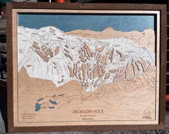 May include: A wooden map of Jackson Hole, Grand Tetons, Wyoming. The map shows the ski runs and terrain of the area. The map is framed in a dark wood frame. The text "JACKSON HOLE" is printed in the lower right corner of the map. The text "Grand Tetons" is printed below "JACKSON HOLE". The text "Wyoming" is printed below "Grand Tetons". The text "slopes" is printed in the lower right corner of the map.