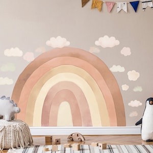 Puede incluir: Un gran adhesivo de pared de arcoíris de acuarela en tonos de rosa, naranja y amarillo con nubes blancas. El adhesivo está en una pared marrón claro en un ambiente de guardería.