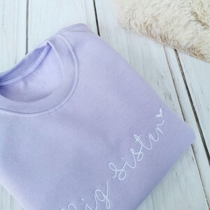 May include: A lavender sweatshirt with the text "Big Sister" embroidered in white with a small heart.
