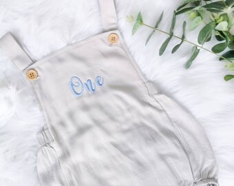 May include: A light gray baby onesie with blue embroidered text that says "One". The onesie has straps and wooden buttons.