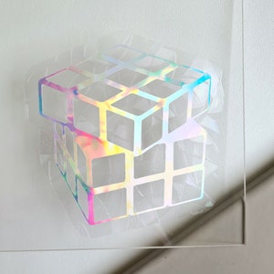 Puede incluir: Una pegatina holográfica de un cubo de Rubik sobre un fondo blanco.