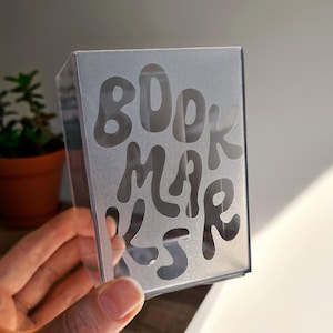 May include: A rectangular, semi-transparent frosted plastic storage box featuring the word BOOKMARKS cut out in a large, wavy, rounded font on the front panel. The container has a clear, smooth finish and a structured, upright design.