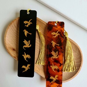 K&ouml;nnte beinhalten: Zwei Lesezeichen mit goldenen Drachen-Silhouetten. Ein Lesezeichen ist schwarz mit goldenen Drachen und das andere ist braun mit goldenen Drachen. Beide Lesezeichen haben goldene Quasten.