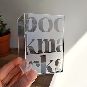 May include: A clear, rectangular acrylic container featuring a frosted, semi-transparent finish. The front surface displays the word "bookmarks" printed in a bold, sans-serif typeface, with the letters stacked vertically across the face.