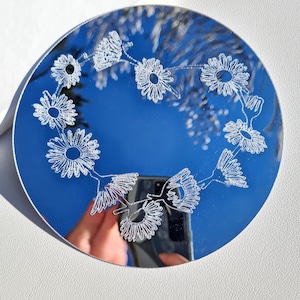 Puede incluir: Un espejo redondo plateado con un diseño floral de margaritas blancas grabado en la superficie. El espejo refleja un cielo azul.