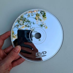 May include: A reflective DVD disc with the word "DVD" printed on it. The disc reflects a blue sky and tree branches. The disc is held by a person.