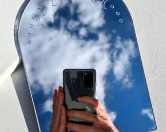 May include: A silver-colored, arched mirror with a crescent moon phase design etched along the top edge. The mirror reflects a blue sky with clouds. The mirror has a rectangular base.