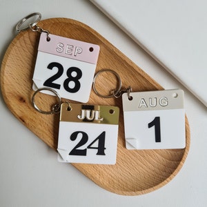 Peut inclure: Trois porte-clés calendrier blancs avec des chiffres noirs et des accents dorés ou roses. Les porte-clés représentent les mois de juillet, août et septembre.