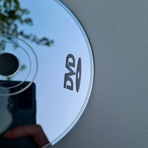 May include: A silver DVD disc with the word "DVD" printed in black. The disc reflects the surrounding environment, including a hand holding a phone. The disc is on a gray surface.