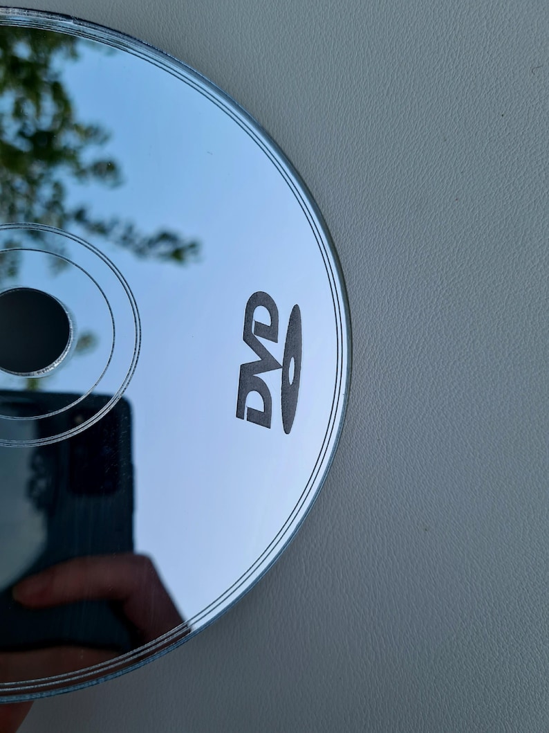 May include: A silver DVD disc with the word "DVD" printed in black. The disc reflects the surrounding environment, including a hand holding a phone. The disc is on a gray surface.