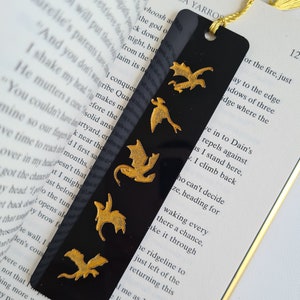 K&ouml;nnte beinhalten: Schwarzes Lesezeichen mit einem goldenen Drachen-Design. Das Lesezeichen hat einen goldenen Quasten und ist aus robustem Material gefertigt.