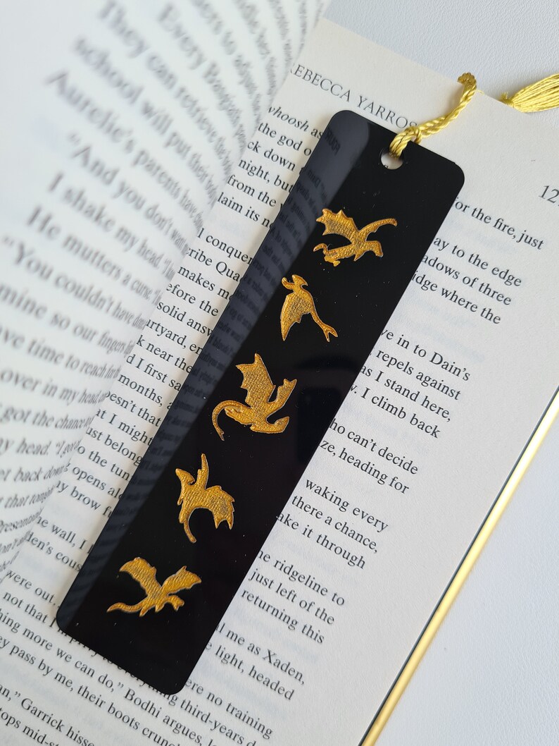 K&ouml;nnte beinhalten: Schwarzes Lesezeichen mit einem goldenen Drachen-Design. Das Lesezeichen hat einen goldenen Quasten und ist aus robustem Material gefertigt.
