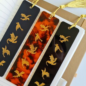 K&ouml;nnte beinhalten: Drei Lesezeichen mit goldenen Drachen-Silhouetten auf schwarzem und orangefarbenem Hintergrund. Die Lesezeichen haben goldene Quasten.