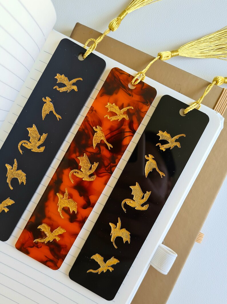 K&ouml;nnte beinhalten: Drei Lesezeichen mit goldenen Drachen-Silhouetten auf schwarzem und orangefarbenem Hintergrund. Die Lesezeichen haben goldene Quasten.