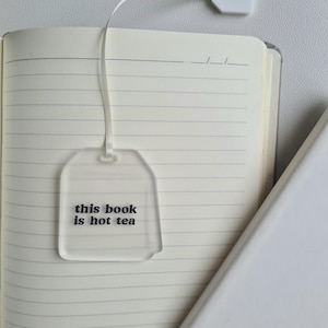 Puede incluir: Un marcap&aacute;ginas transparente de acr&iacute;lico con forma de bolsita de t&eacute;, con el texto "this book is hot tea" en negro. El marcap&aacute;ginas est&aacute; unido a una cinta blanca y a una peque&ntilde;a etiqueta blanca, descansando sobre las p&aacute;ginas abiertas de un cuaderno rayado.