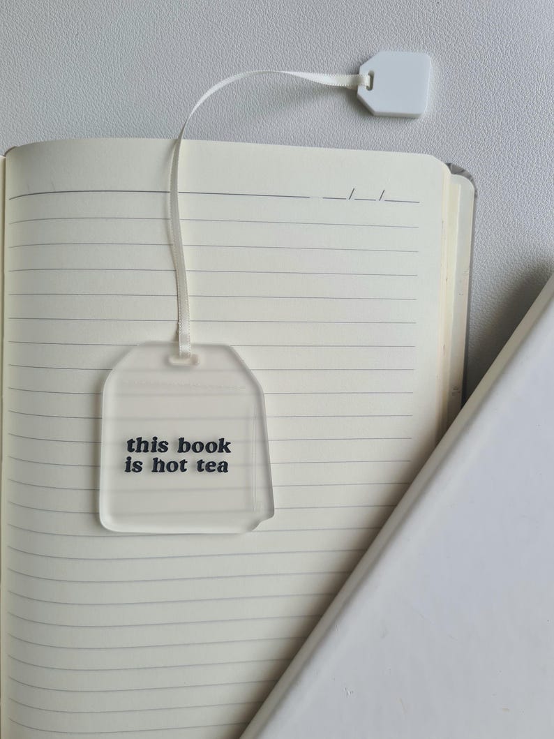 Puede incluir: Un marcap&aacute;ginas transparente de acr&iacute;lico con forma de bolsita de t&eacute;, con el texto "this book is hot tea" en negro. El marcap&aacute;ginas est&aacute; unido a una cinta blanca y a una peque&ntilde;a etiqueta blanca, descansando sobre las p&aacute;ginas abiertas de un cuaderno rayado.