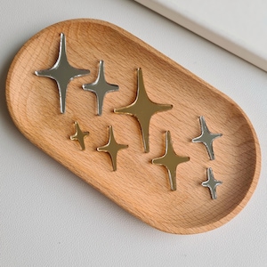 Op de afbeelding: Een assortiment stervormige bedels in verschillende maten en metallic afwerkingen, waaronder zilver en goud, is gerangschikt op een houten dienblad. De bedels hebben vier punten en worden gepresenteerd tegen een neutrale achtergrond.