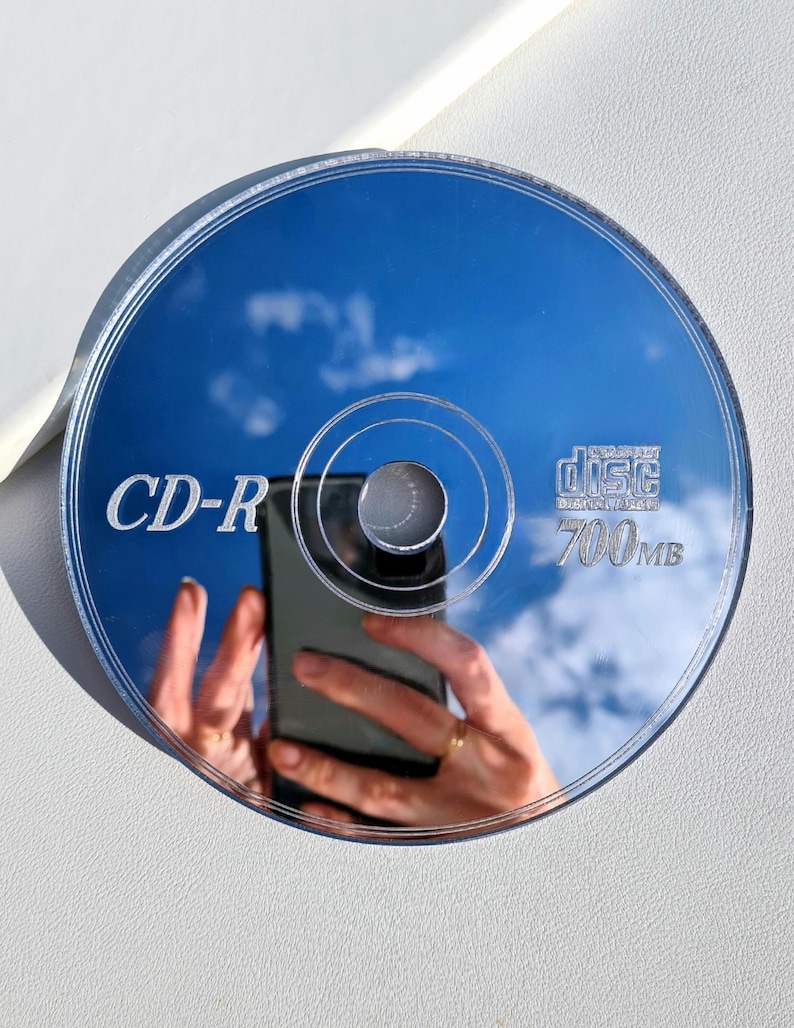 May include: A reflective CD-R disc with the text "CD-R" and "700MB" printed on it. The disc reflects a blue sky with clouds and a person holding a phone. The disc is circular and has a silver center.