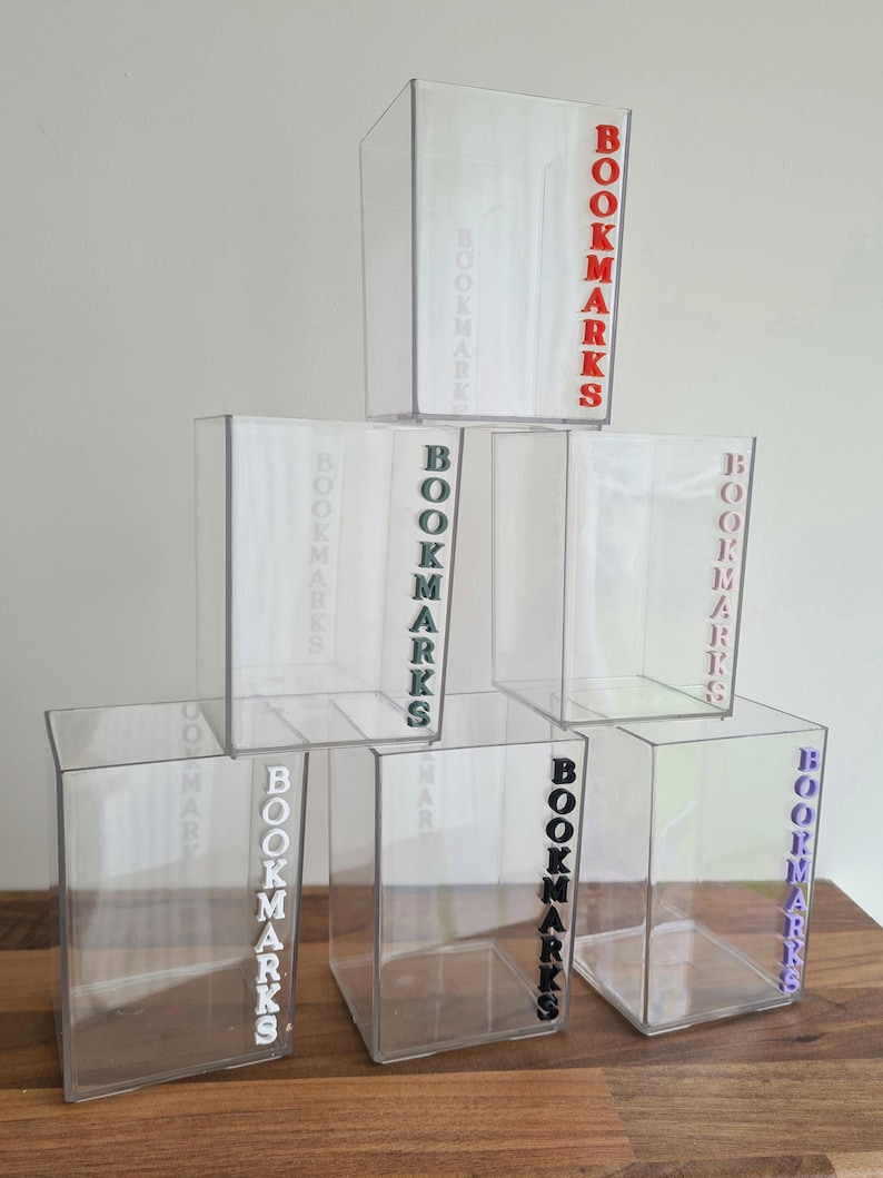 May include: Six clear acrylic containers with the word "BOOKMARKS" printed on the side in different colors. The colors are red, green, white, black, purple, and light blue.