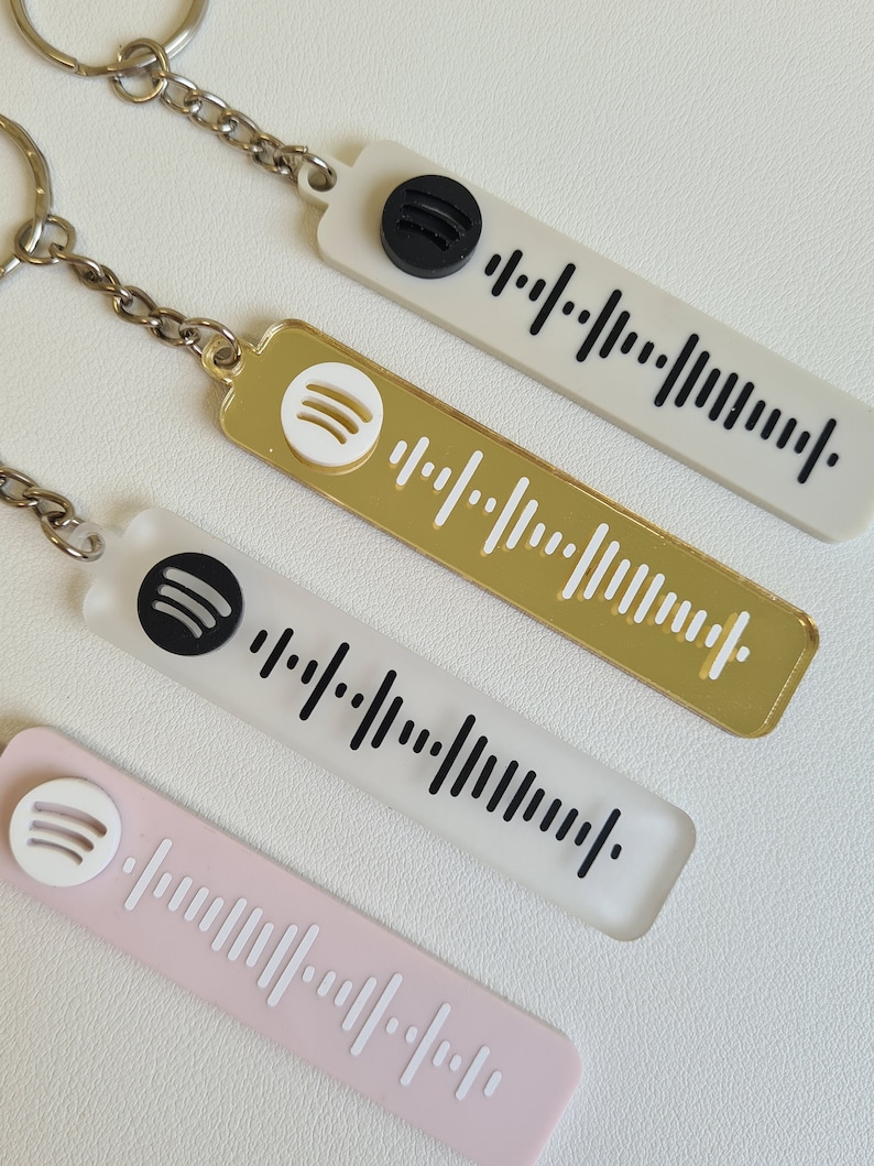 Puede incluir: Cuatro llaveros con dise&ntilde;os de c&oacute;digo Spotify. Los llaveros son blancos, dorados y rosas, con patrones de barras en blanco y negro. Cada llavero tiene un logotipo de Spotify y una cadena de metal.