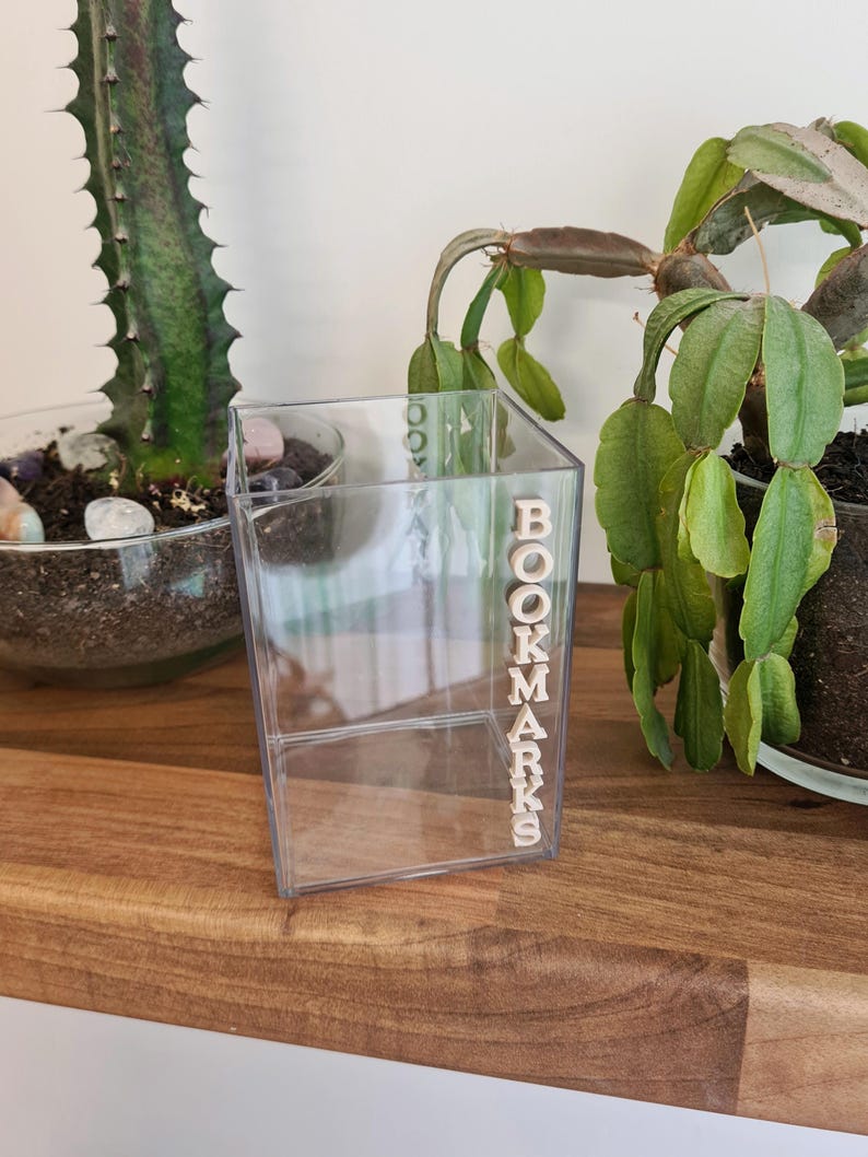 May include: A clear plastic container with the word "BOOKMARKS" spelled out in wooden letters. The container is sitting on a wooden surface.