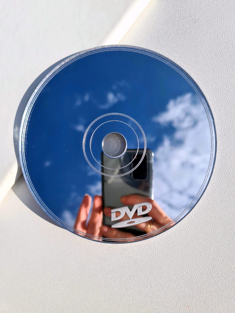 May include: A reflective DVD disc with a blue sky and clouds reflected on its surface. The word "DVD" is visible on the disc. The disc is circular with concentric rings and a silver center.
