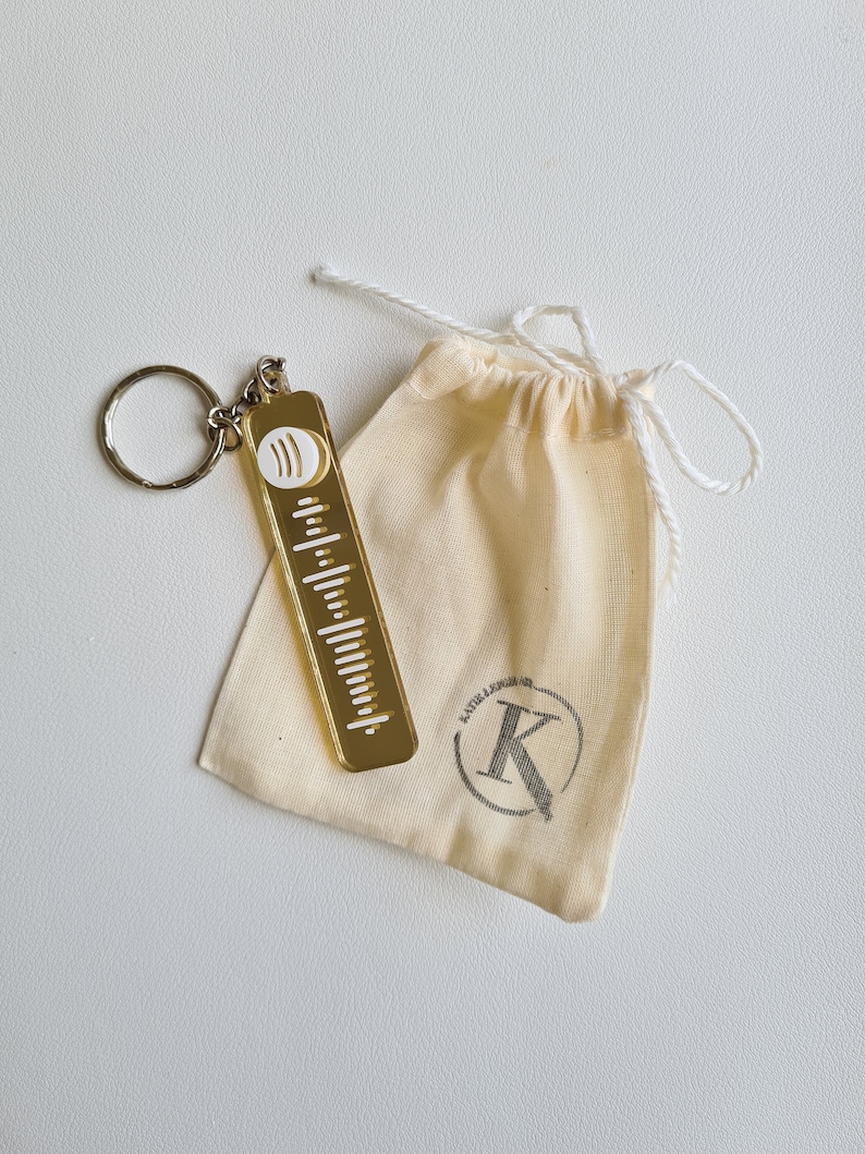 Puede incluir: Un llavero dorado con un dise&ntilde;o de c&oacute;digo Spotify y un llavero plateado. El llavero est&aacute; junto a una peque&ntilde;a bolsa con cord&oacute;n de color crema con un logotipo impreso. La bolsa est&aacute; hecha de una tela suave.
