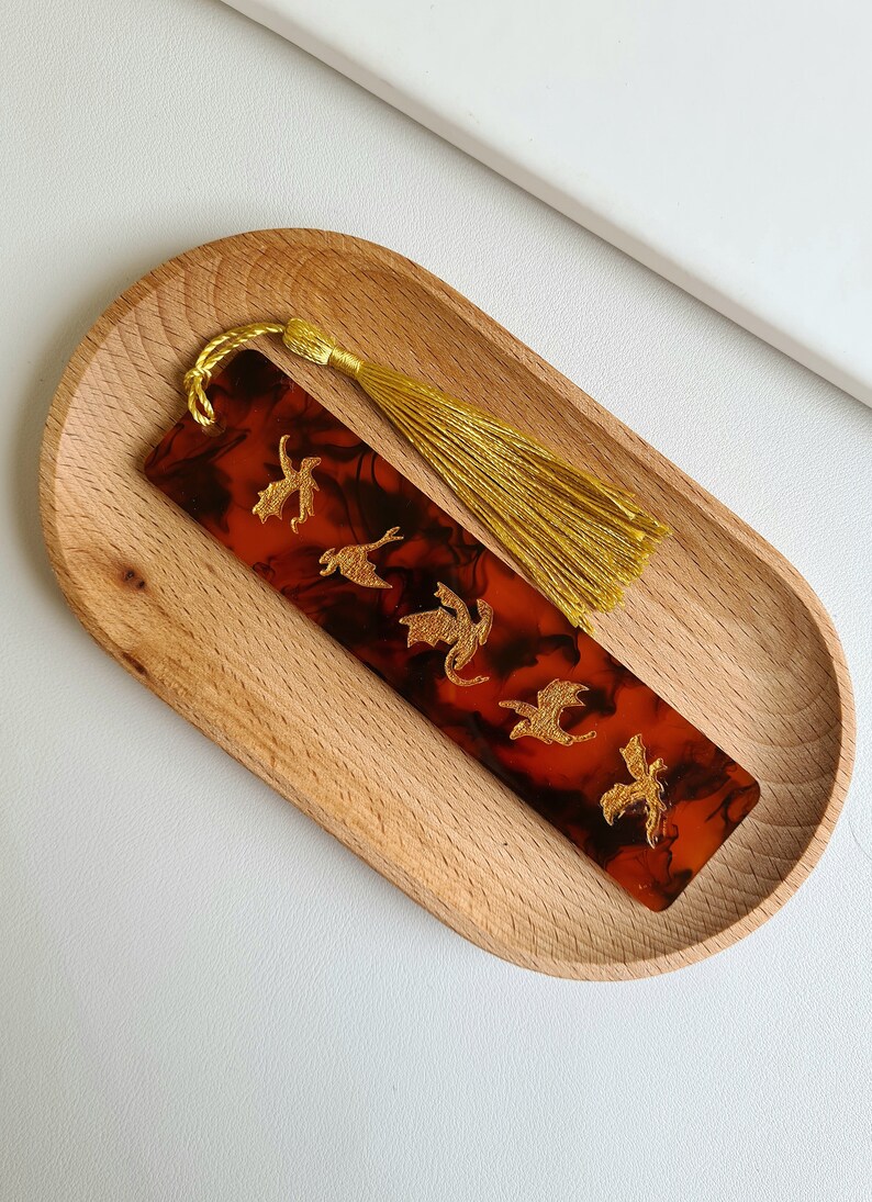 K&ouml;nnte beinhalten: Ein Lesezeichen aus rotem und schwarzem Harz mit einem goldenen Quasten und einem Design aus f&uuml;nf goldenen Drachen.