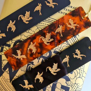 K&ouml;nnte beinhalten: Vier schwarze und braune Lesezeichen mit goldenen Drachenmotiven. Die Lesezeichen sind aus einem durchsichtigen Material gefertigt und haben einen Quasten.