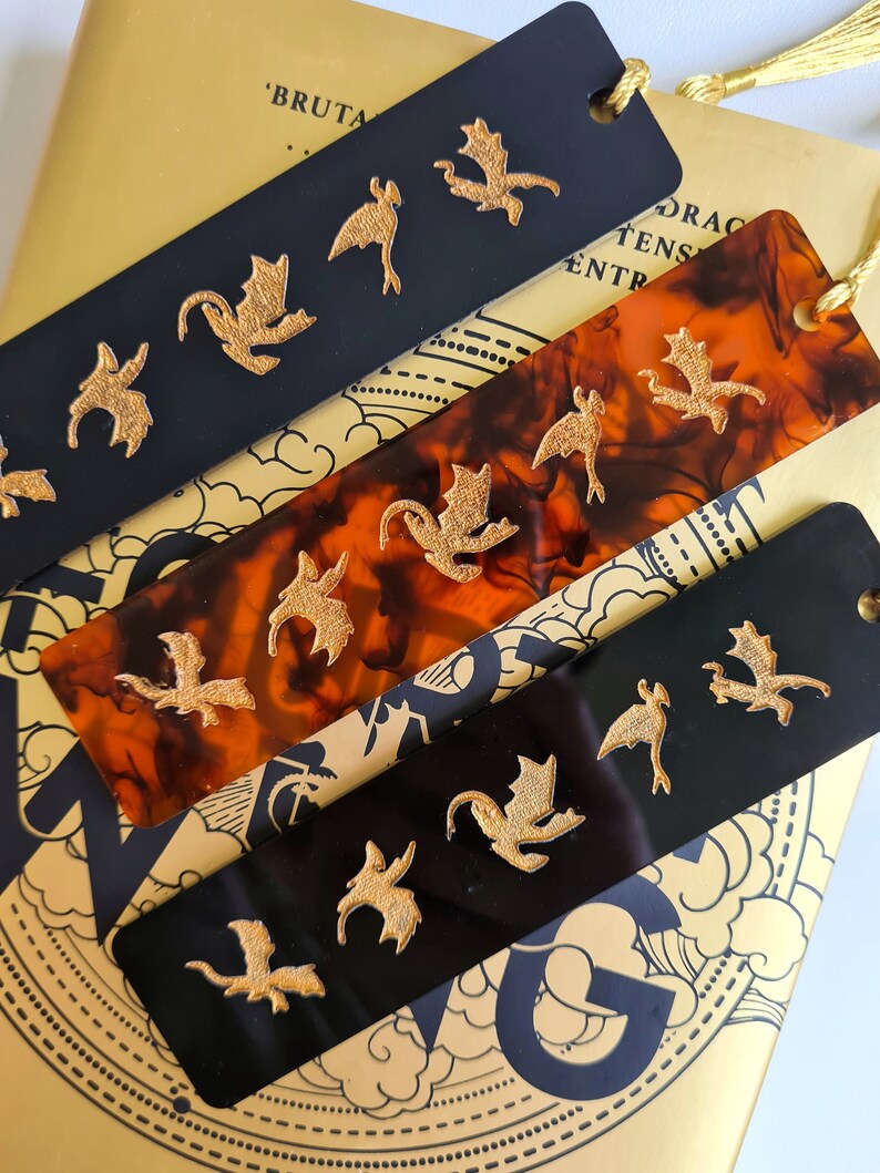 K&ouml;nnte beinhalten: Vier schwarze und braune Lesezeichen mit goldenen Drachenmotiven. Die Lesezeichen sind aus einem durchsichtigen Material gefertigt und haben einen Quasten.