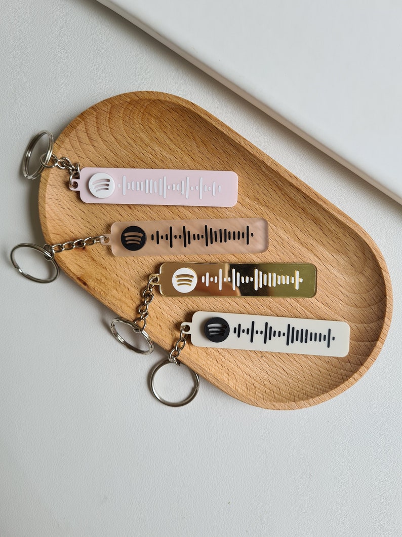 Puede incluir: Cuatro llaveros con dise&ntilde;os de c&oacute;digo Spotify en varios colores: rosa claro, transparente, verde oliva y blanco. Cada llavero tiene una cadena plateada y un logotipo de Spotify. Los llaveros se muestran en una bandeja de madera.
