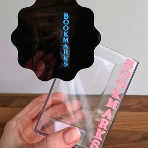 Puede incluir: Un soporte transparente para marcapáginas de plástico con la palabra "BOOKMARKS" en letras rosas en el lateral. Una forma negra con bordes festoneados con la palabra "BOOKMARKS" en letras azules está encima del soporte.
