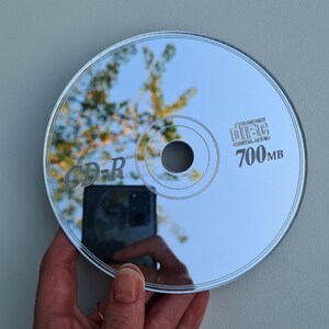 May include: A reflective CD-R disc with the text "CD-R" and "700MB" printed on the surface. The disc reflects the surrounding environment, including a hand holding the disc and a view of the sky and trees.