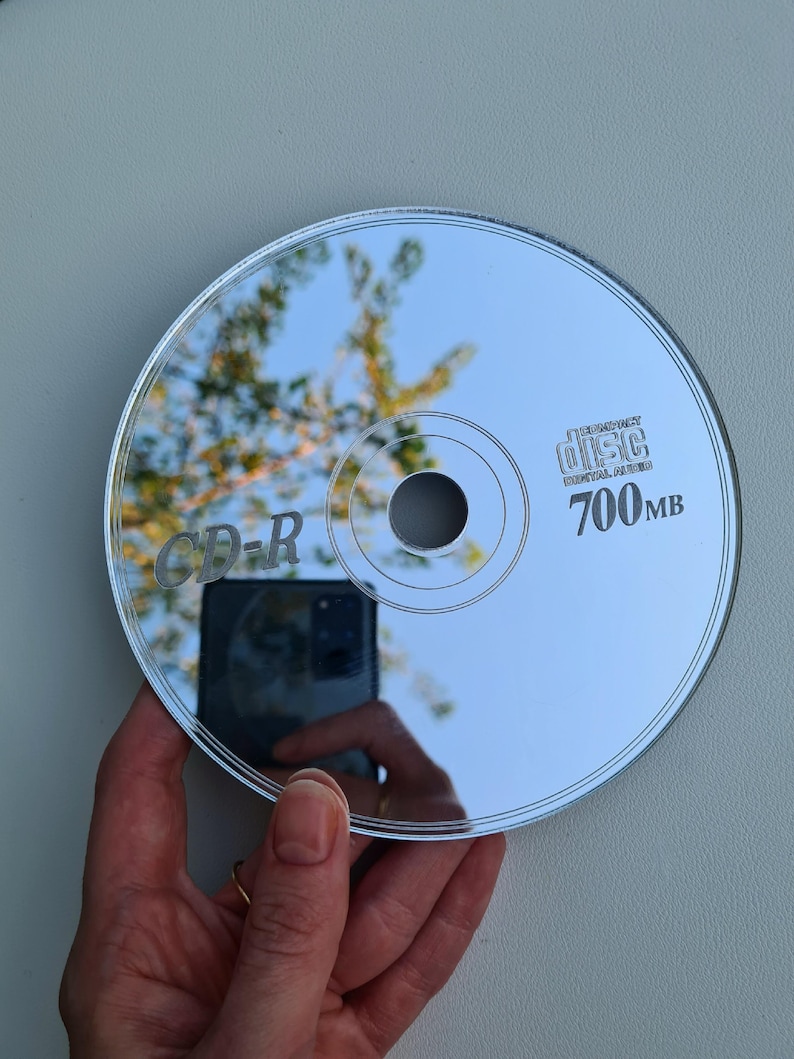 May include: A reflective CD-R disc with the text "CD-R" and "700MB" printed on the surface. The disc reflects the surrounding environment, including a hand holding the disc and a view of the sky and trees.