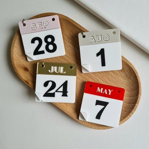 May include: Four small calendar-shaped ornaments with different months and dates. The ornaments are made of white plastic with black numbers and are on a wooden surface. The months are September, August, July, and May. The dates are 28, 1, 24, and 7.