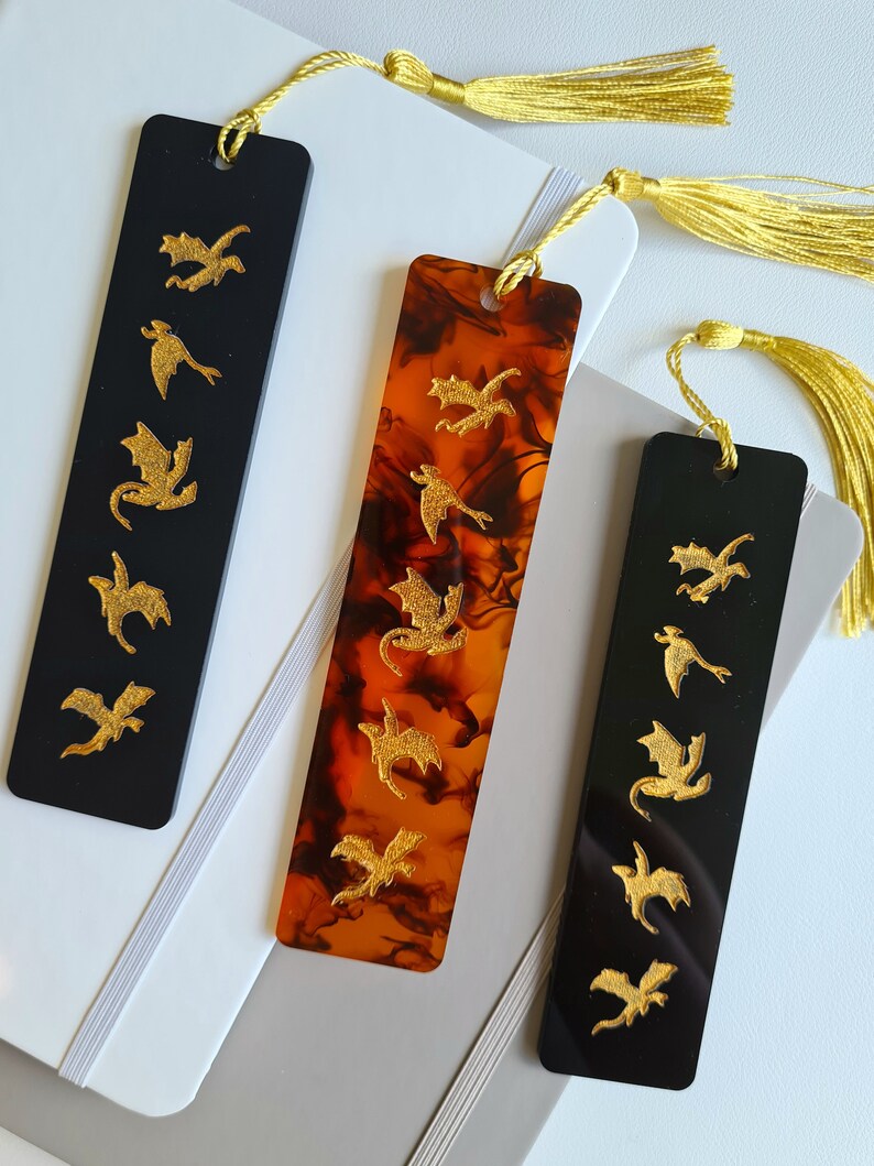 K&ouml;nnte beinhalten: Zwei Lesezeichen mit goldenen Drachenmotiven. Ein Lesezeichen ist schwarz mit goldenen Drachen und das andere ist braun mit goldenen Drachen.