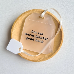 Puede incluir: Un posavasos de madera marr&oacute;n claro sostiene una etiqueta transl&uacute;cida en forma de bolsita de t&eacute; con las palabras "hot tea, warm blanket, good book" impresas en negro. Se adjuntan una cinta y una etiqueta blancas. El posavasos est&aacute; sobre una superficie blanca.